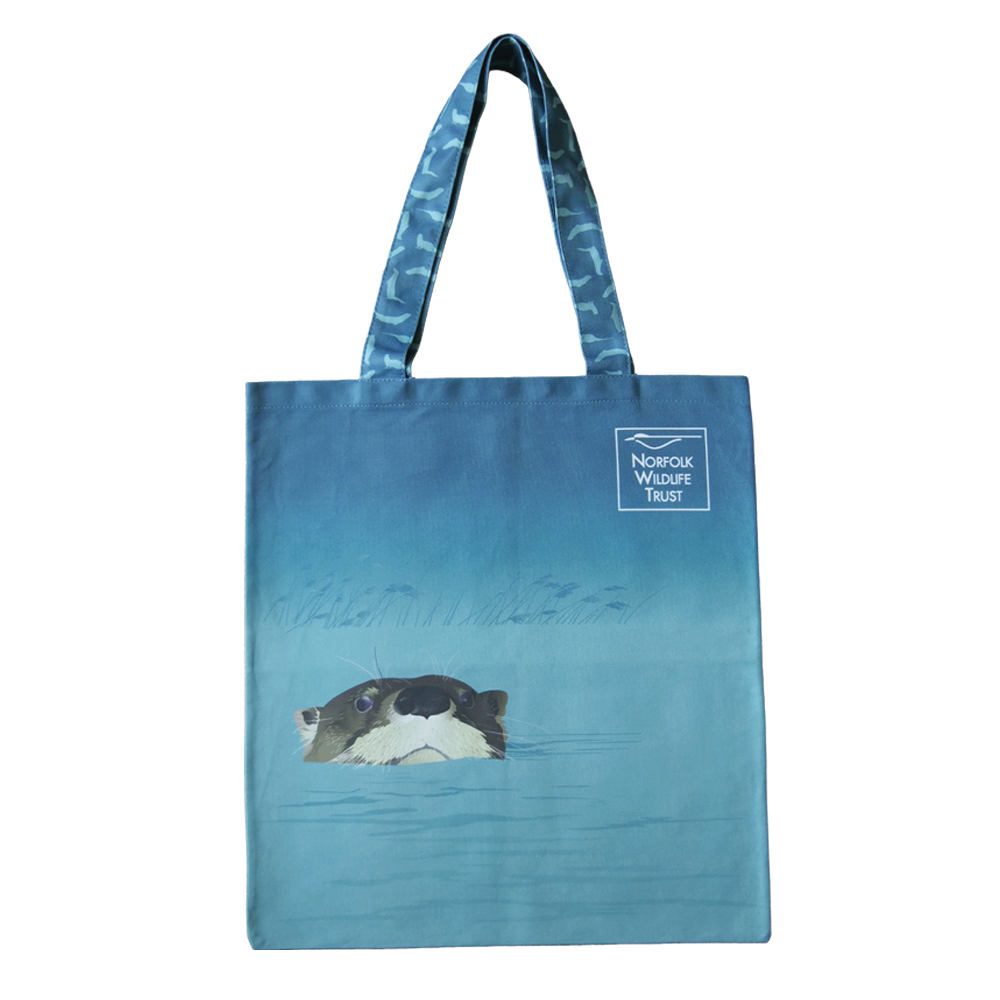 A blue rectangular tote bag features an otter peeking out from a reed-lined river or broad